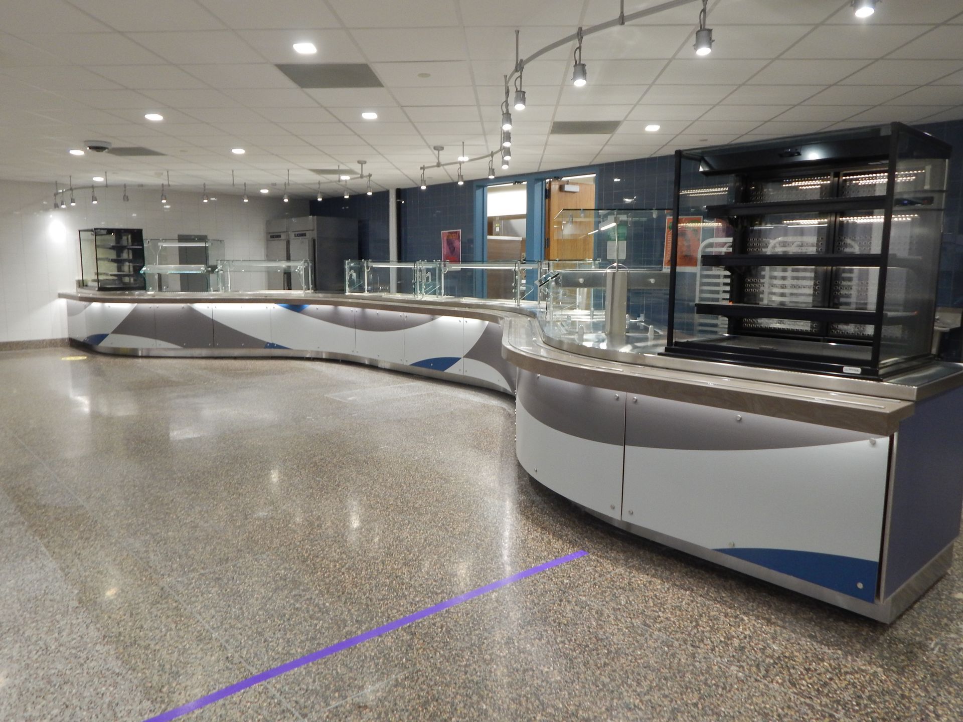 An empty cafeteria serving line with a curved counter, stainless steel fixtures, and a glass-enclosed display case.