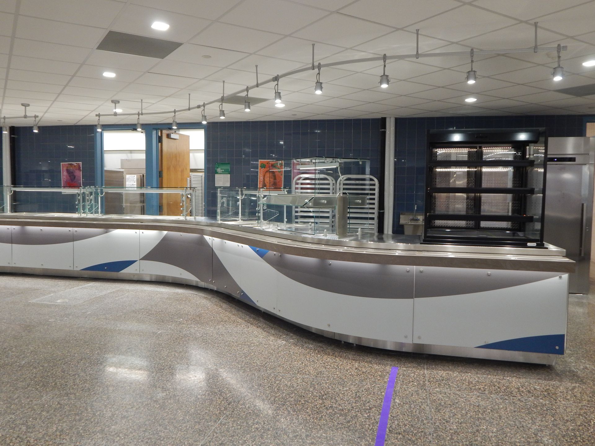 A curved, modern cafeteria serving counter with white, gray, and blue wave-pattern panels in a tiled-floor room.