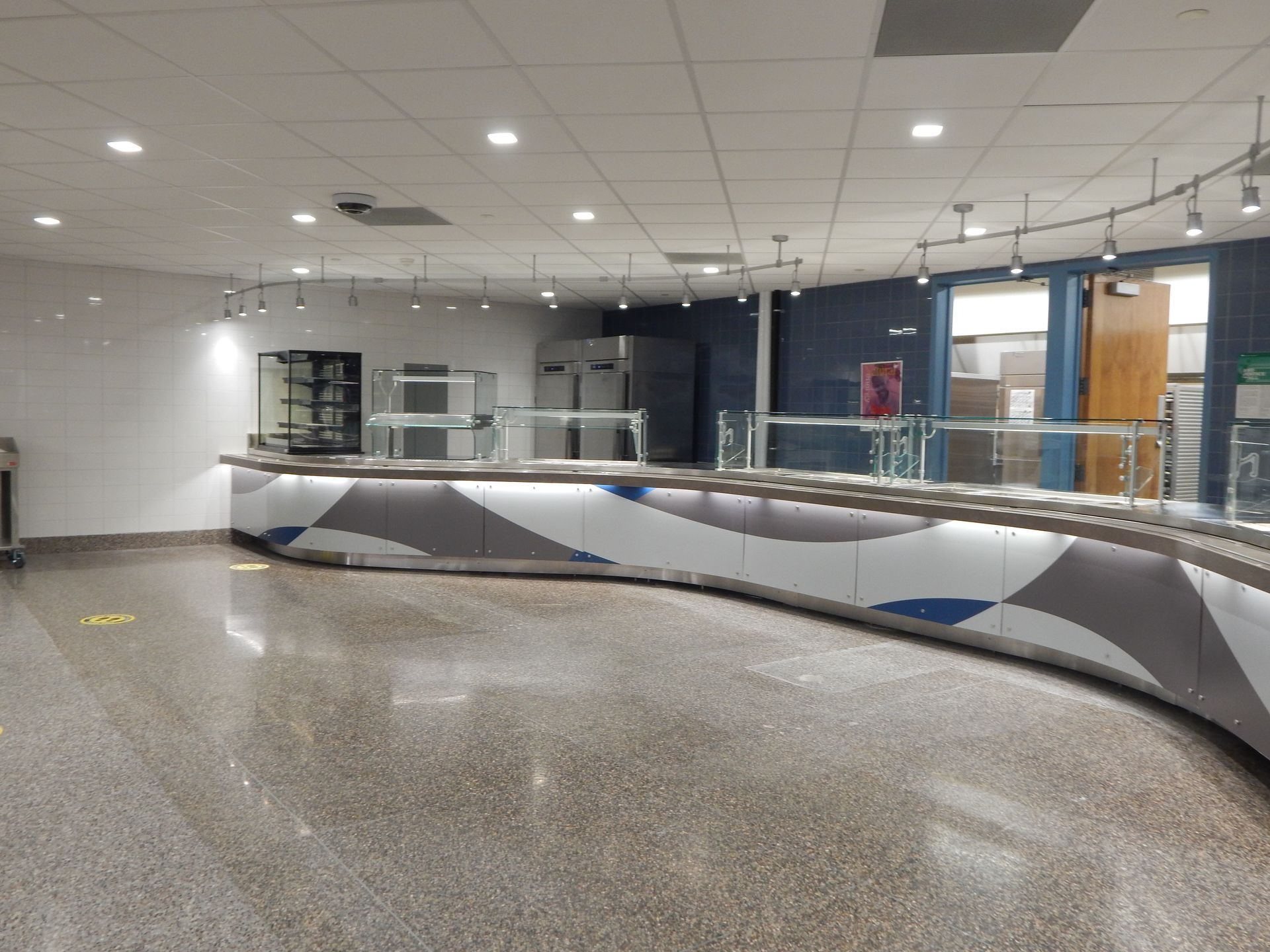 A curved, modern cafeteria serving counter with a white, gray, and blue patterned base and a polished speckled floor.