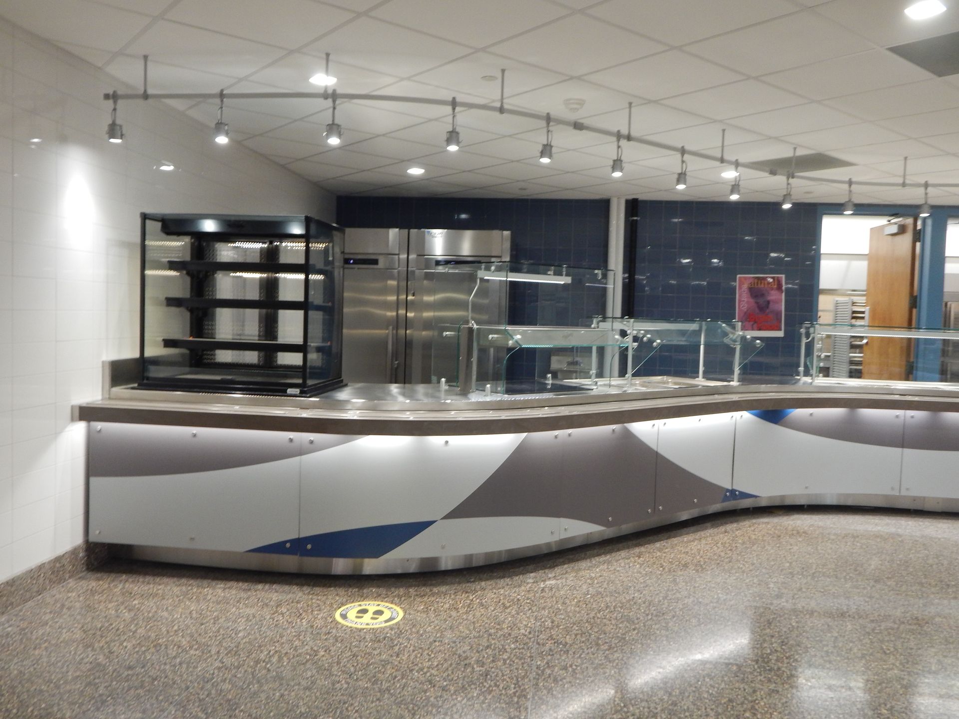 A curved cafeteria serving counter with a multi-shelf display, stainless steel refrigerators, and overhead track lighting.