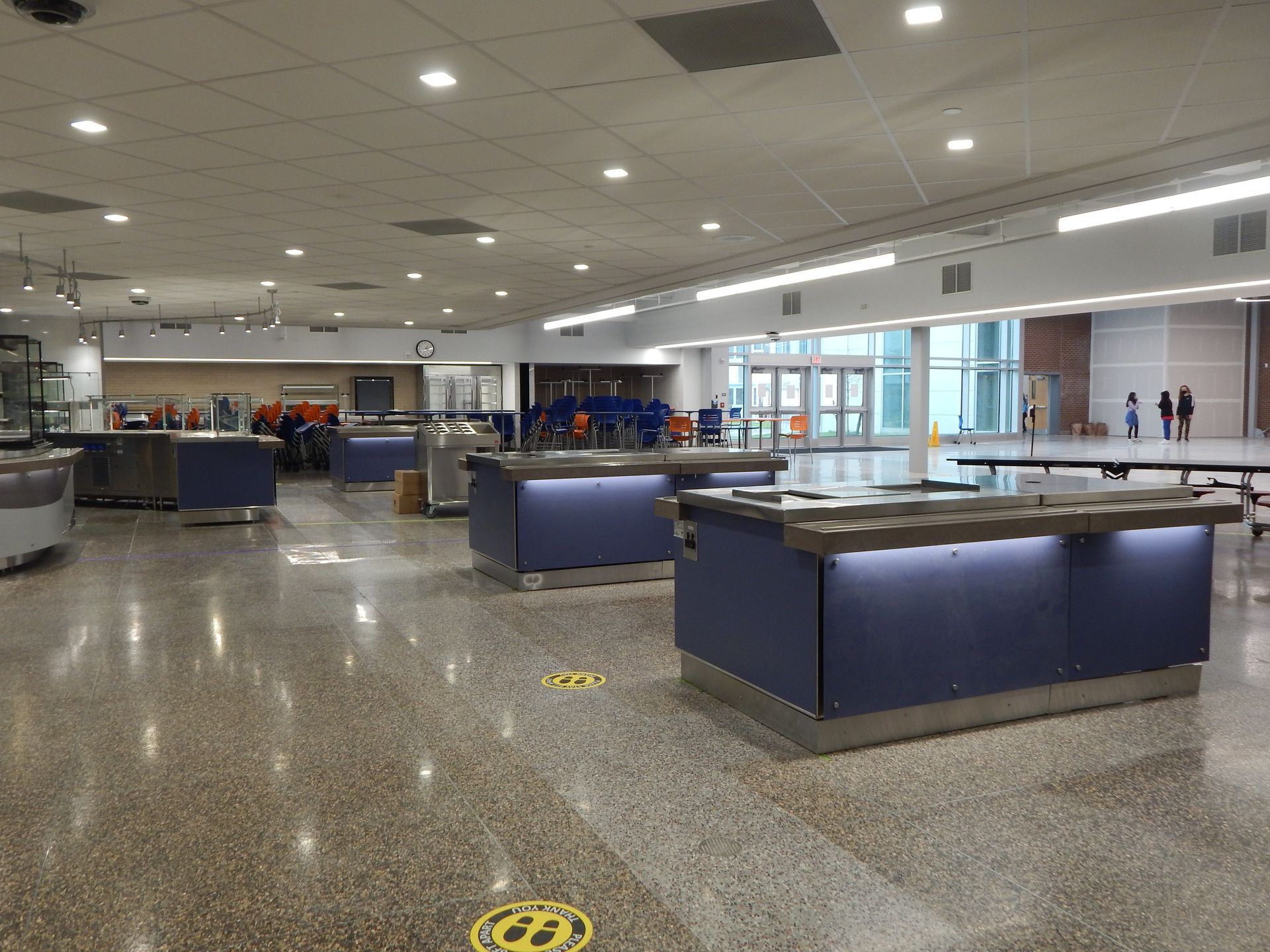 An empty dining hall featuring blue serving counters on a speckled floor and floor-to-ceiling windows in the distance.
