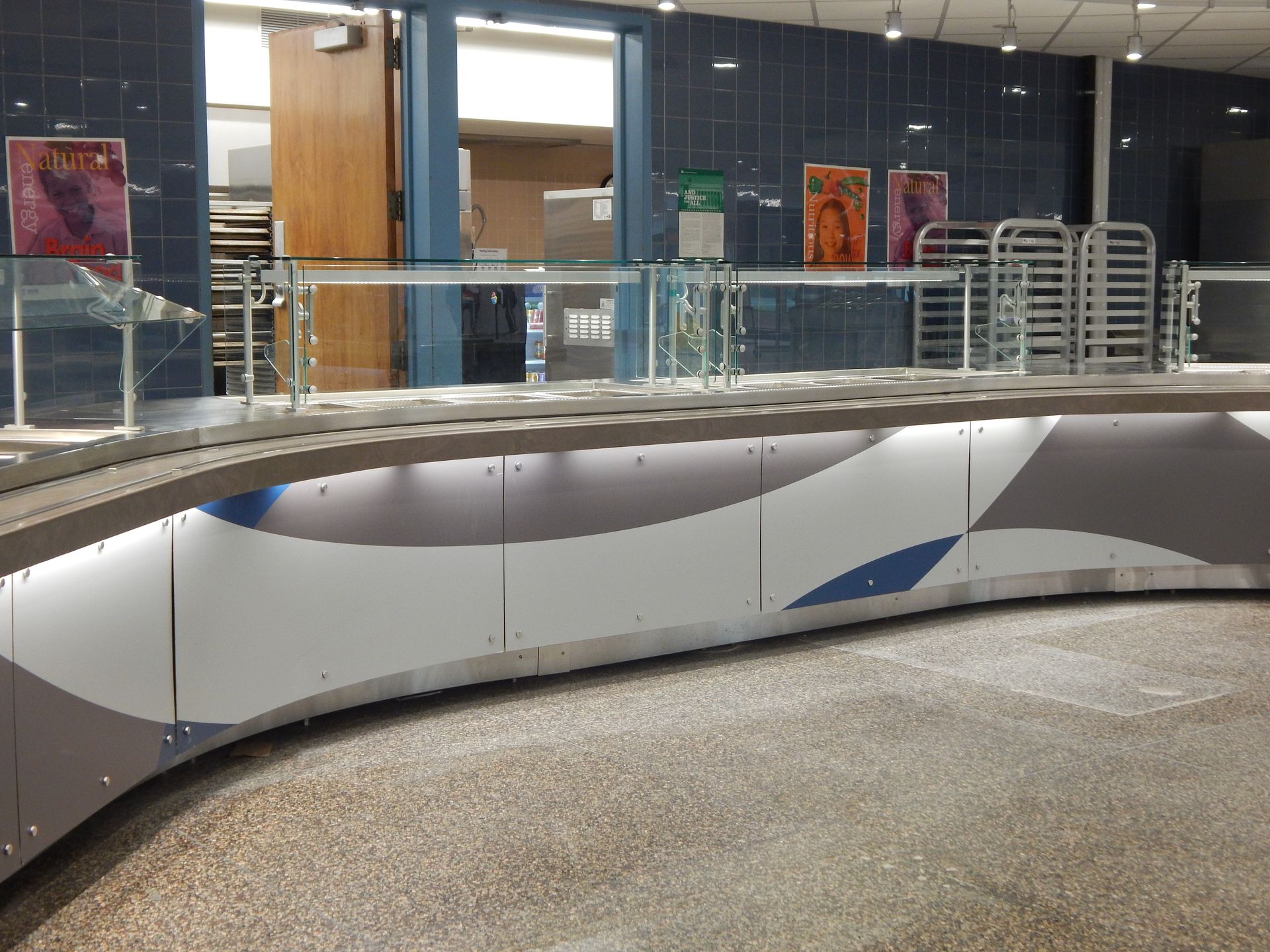 A curved, modern cafeteria serving counter with glass sneeze guards and blue, gray, and white patterned panels.