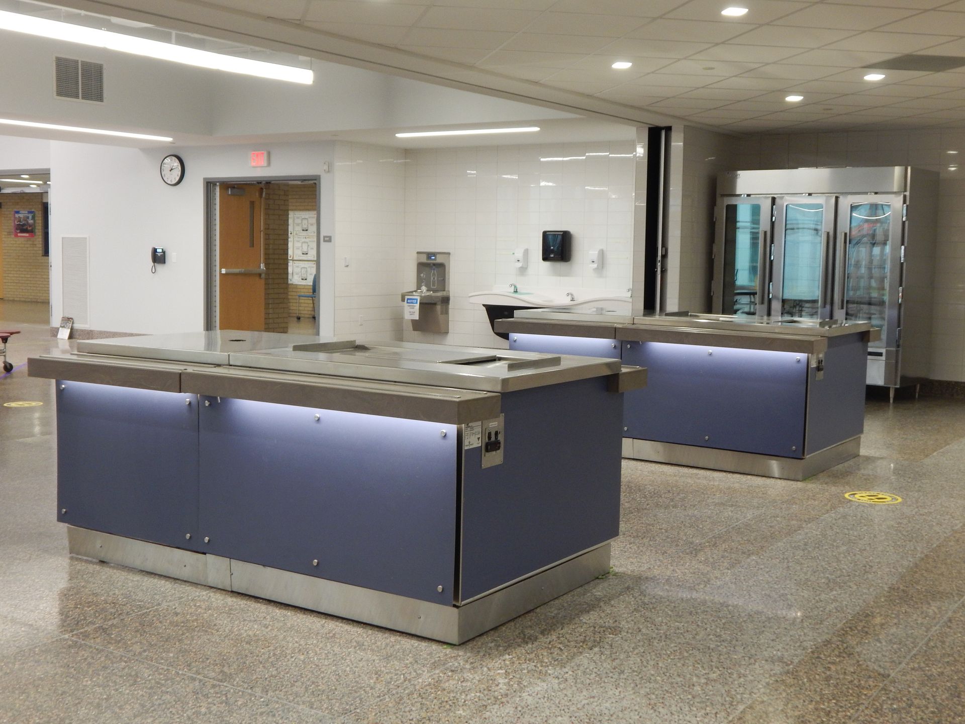 Two blue serving stations with under-counter lighting are positioned in a brightly lit, commercial dining hall.