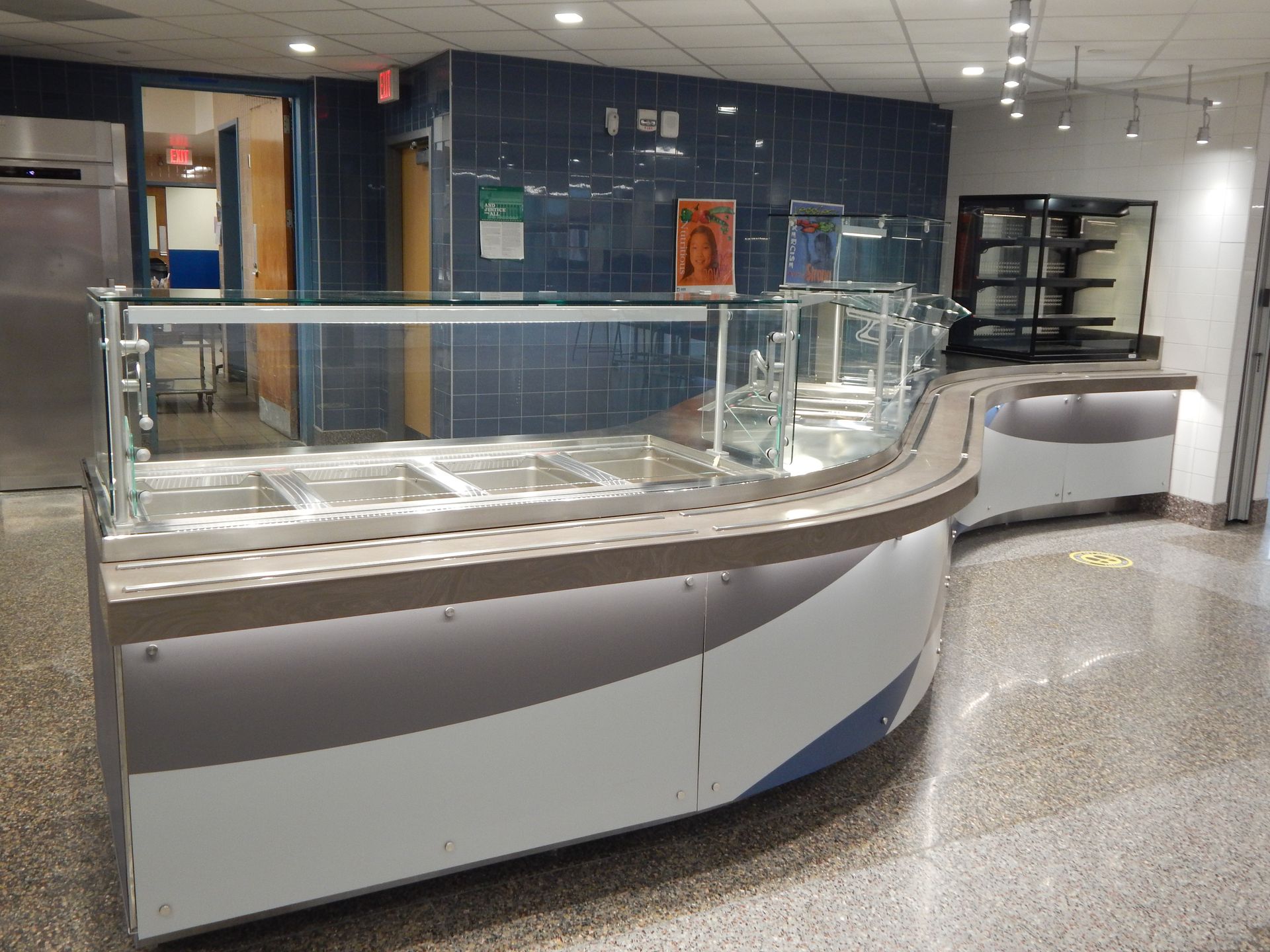 A curved, stainless steel food service counter with glass sneeze guards in a tiled cafeteria.