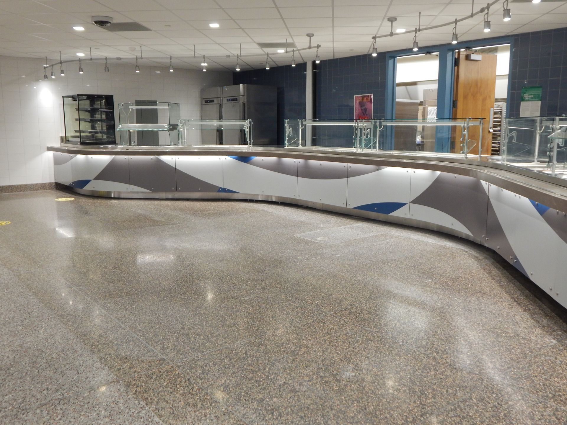 A curved, modern cafeteria service counter with a patterned base and glass sneeze guards in a bright, tiled room.