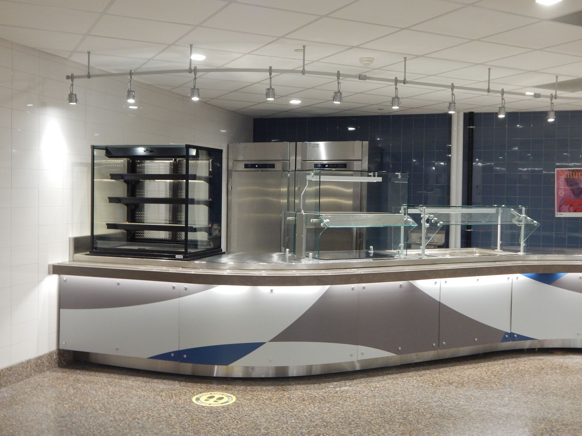 A curved cafeteria serving counter with a refrigerated display case, stainless steel fridges, and overhead lighting.