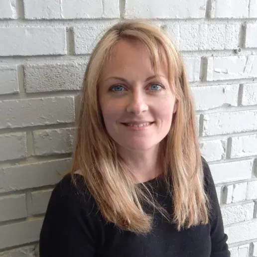 A person with long blonde hair and a neutral expression, posing in front of a white painted brick wall.