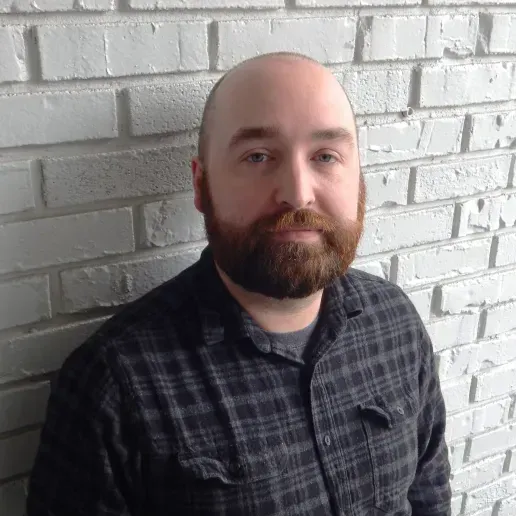 A person with a beard and a plaid shirt stands in front of a white painted brick wall.