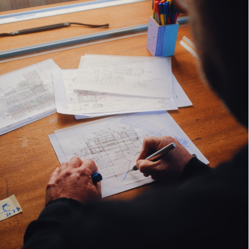 A person in a black sweater draws with a pen on architectural blueprints spread across a wooden drafting table.