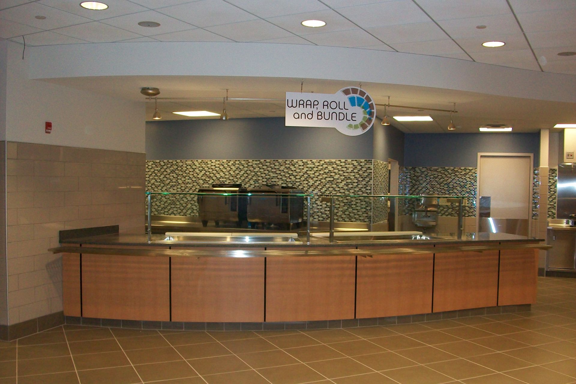 An empty food serving station in a cafeteria with light wood cabinets, tiled walls, and a hanging sign.
