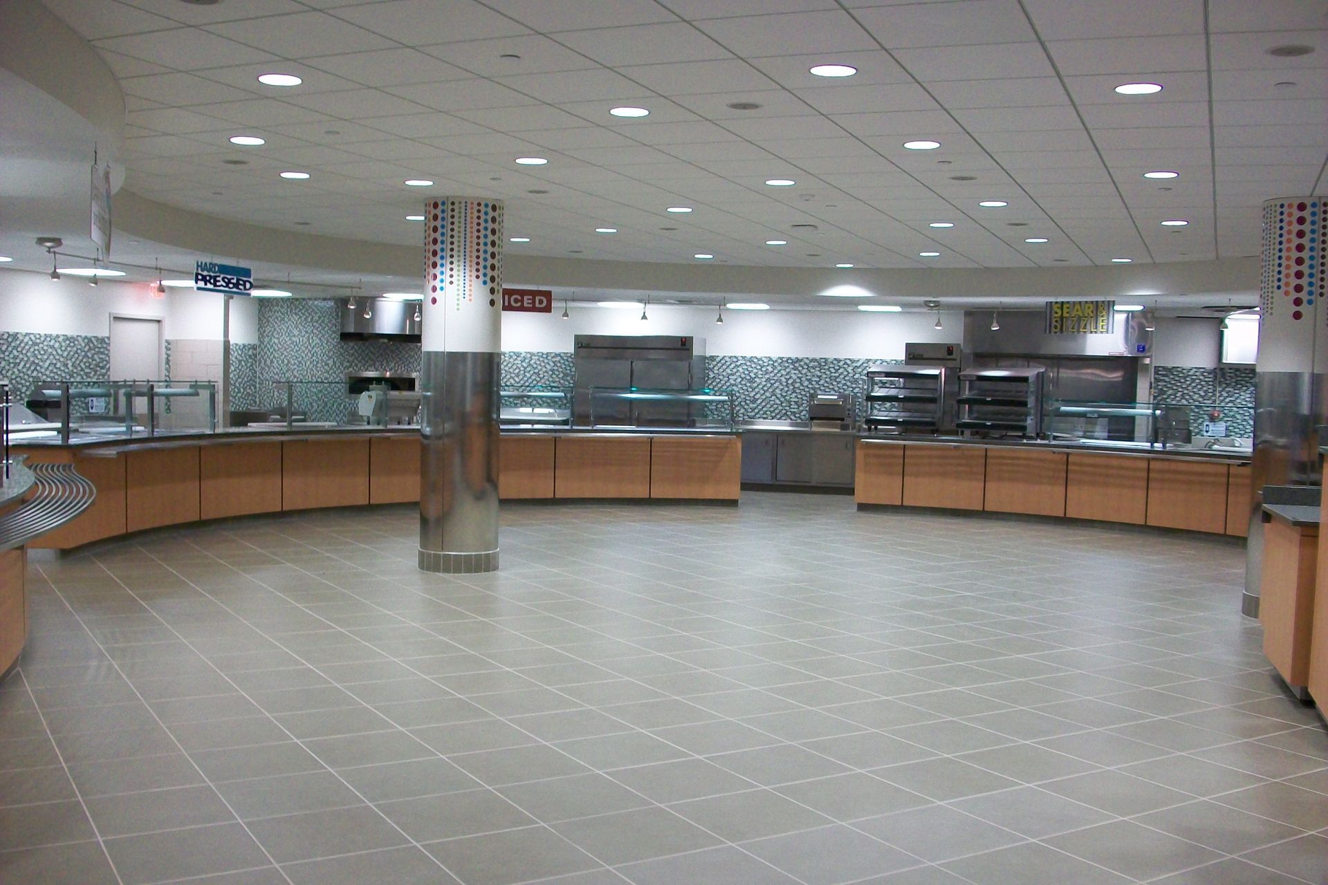 A modern, curved cafeteria serving line featuring light wood cabinets, stainless steel pillars, and a tiled floor.