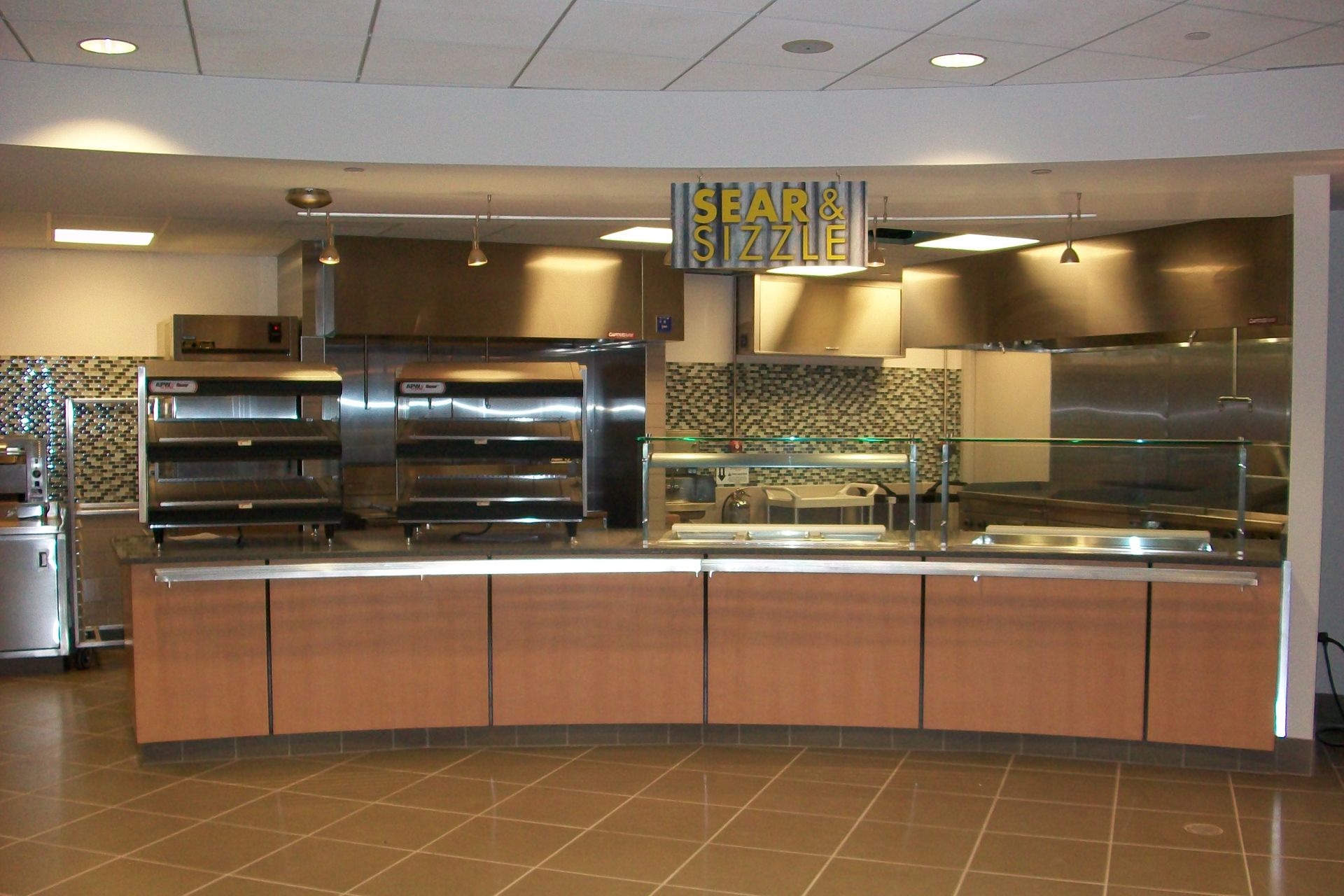 A food service station with a long, light-wood counter and glass sneeze guards in a tiled, well-lit cafeteria setting.