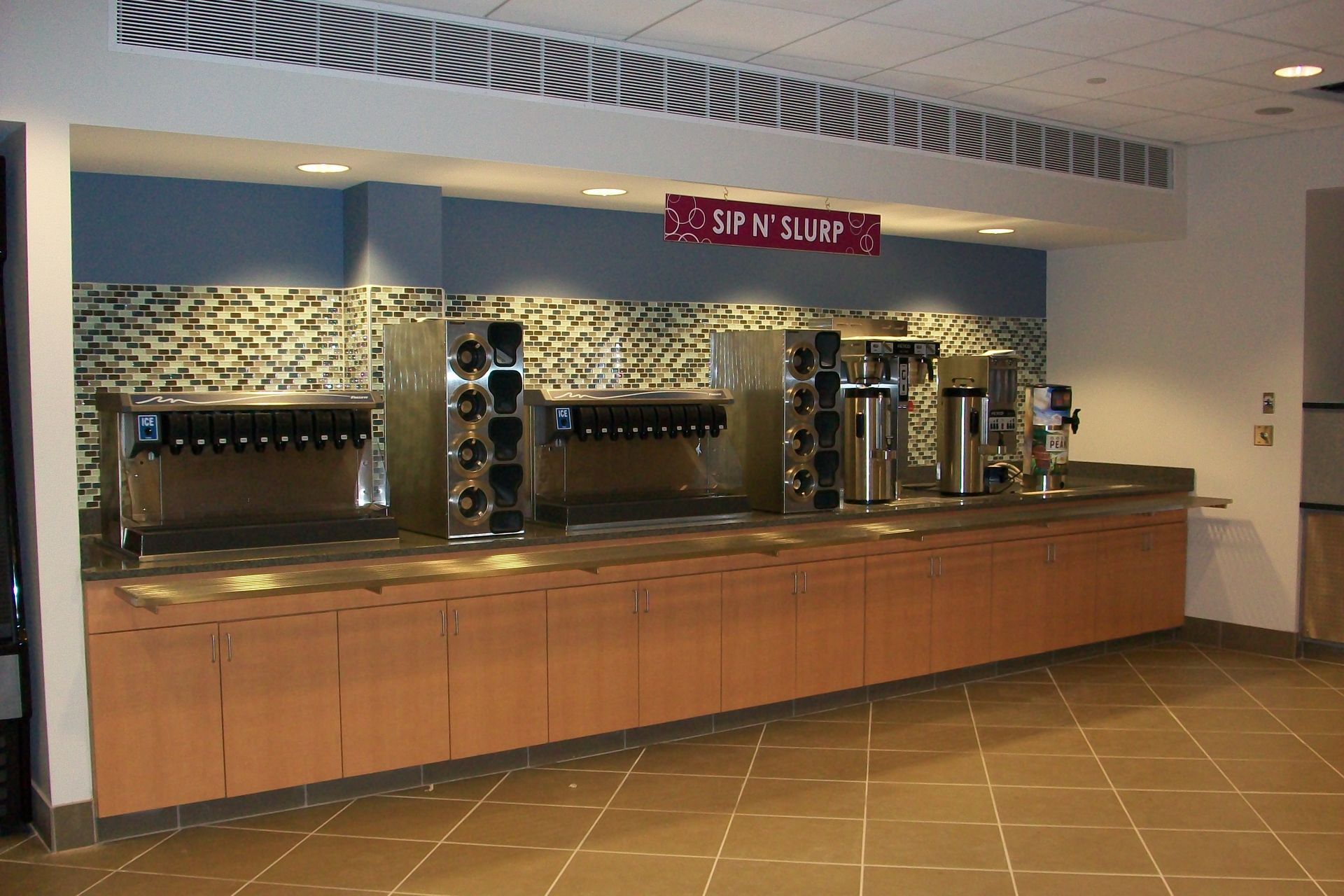 A beverage station with multiple soda dispensers and drink canisters set atop a long wood cabinet against a blue wall.