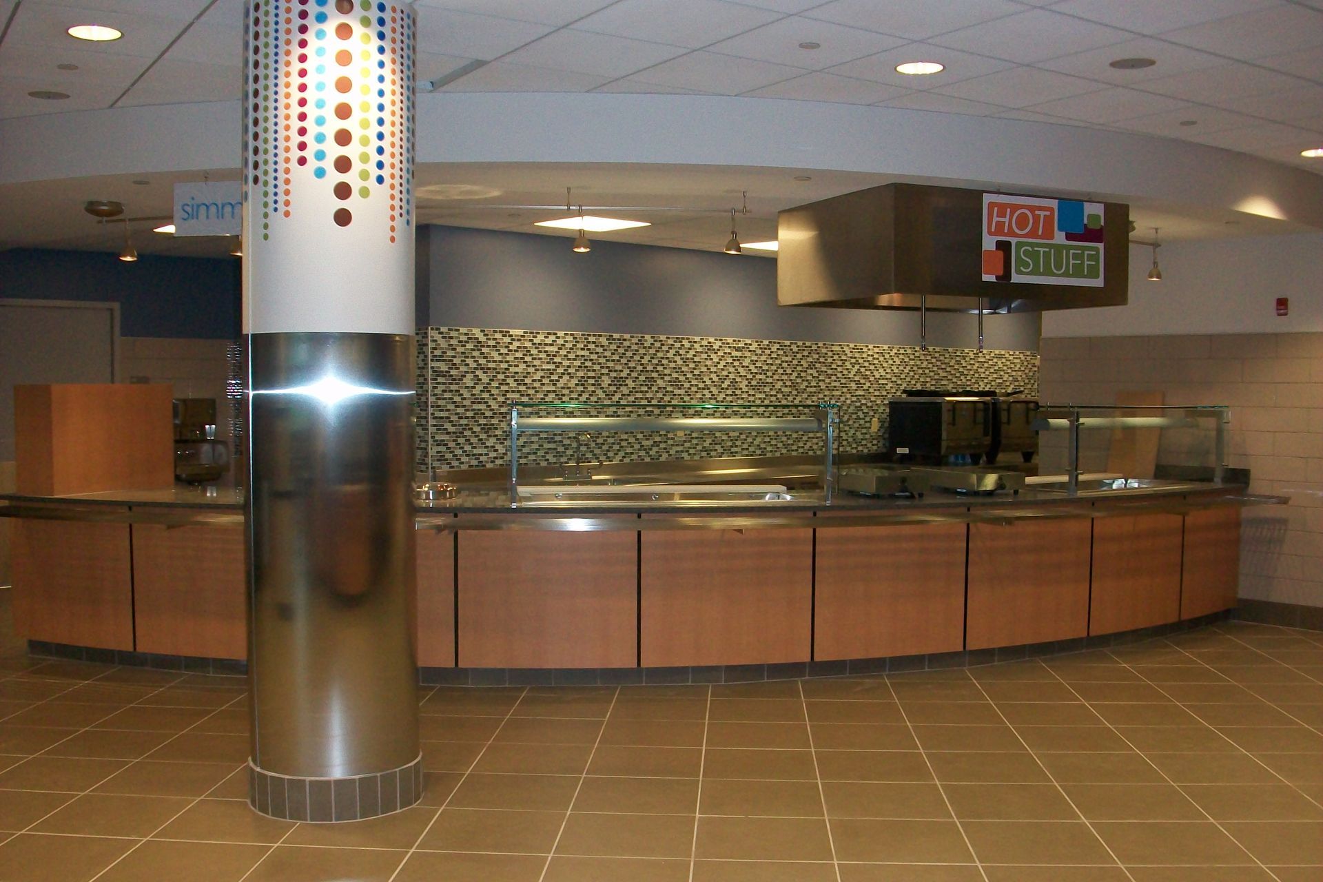 A cafeteria serving station with a curved wood-panel counter, a silver pillar with colorful accents, and a tile backsplash.