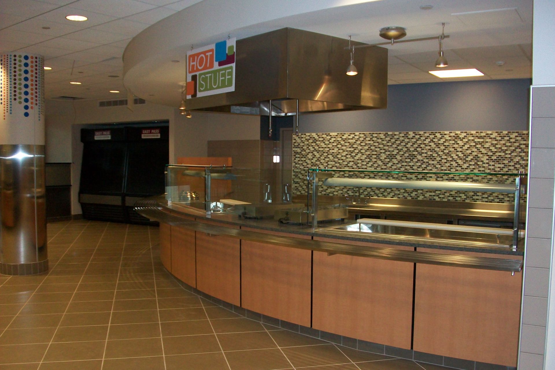 A service counter with a metal hood and glass sneeze guards in a modern dining area with tiled floors and a textured wall.