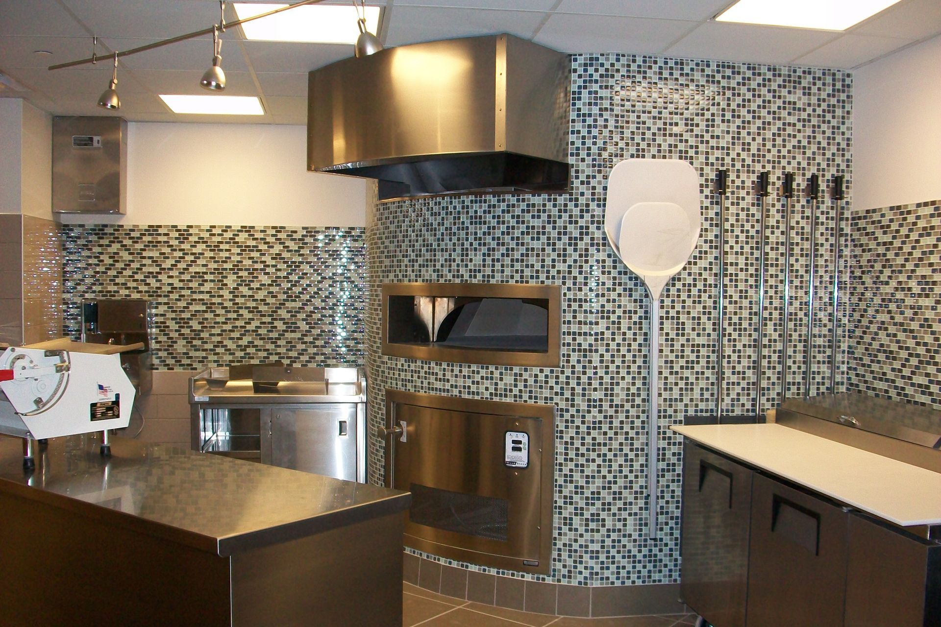 A stainless steel pizza oven and prep station in a modern kitchen with patterned mosaic tile walls.