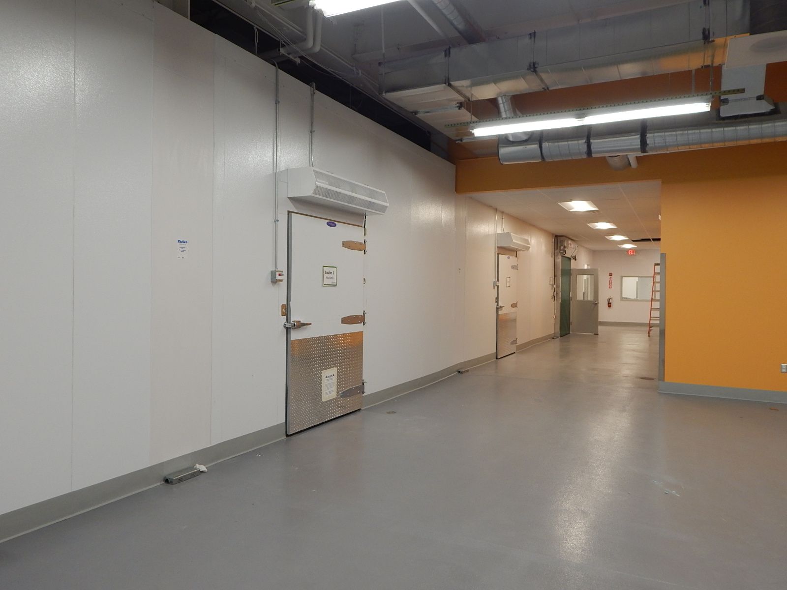 A hallway with a white wall, a metal-paneled freezer door, grey concrete floor, and orange accents on the far wall.
