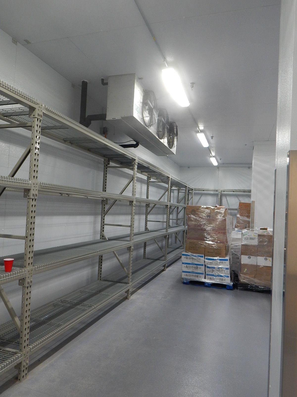 A walk-in refrigerator featuring tall metal shelving units along the walls and pallets of supplies on the floor.