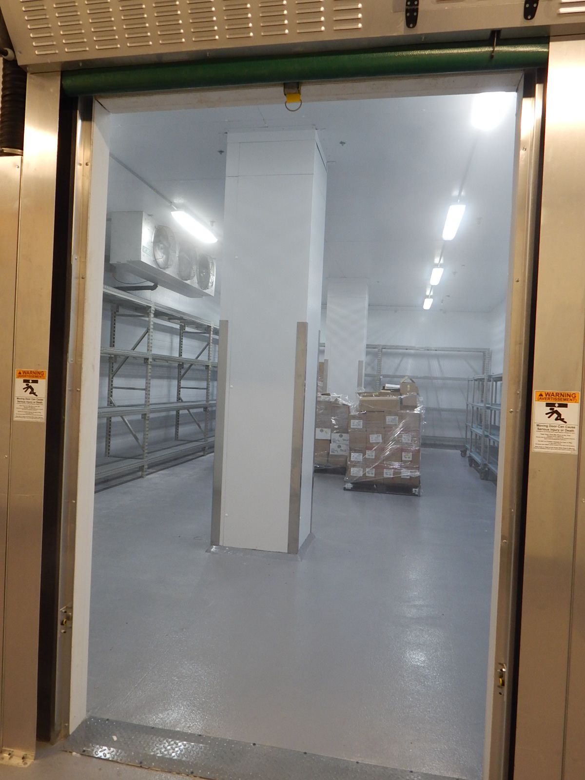 A view through a metal doorway into a brightly lit walk-in freezer with industrial shelving and stacked pallet goods.