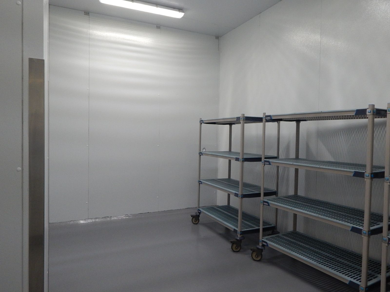 Empty industrial walk-in cooler or storage room with white walls, gray floors, and two metal shelving units.