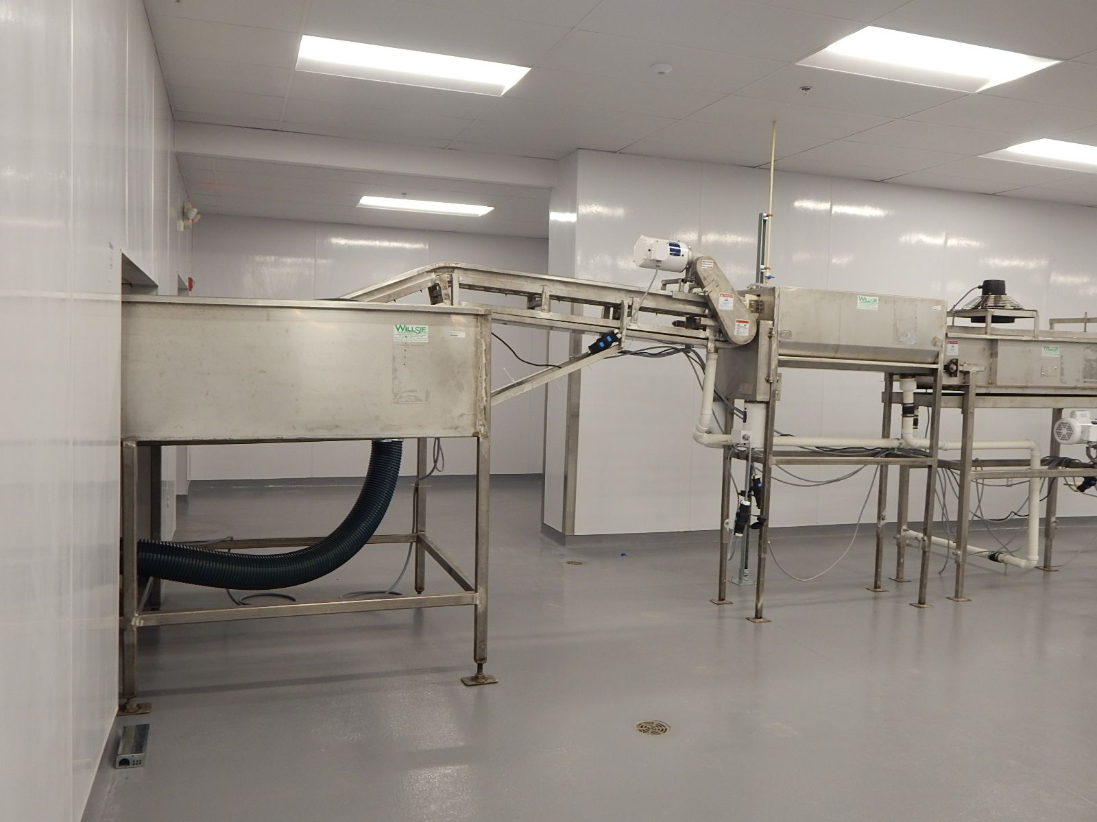 A stainless steel industrial food processing conveyor system in a clean, white-walled commercial facility.