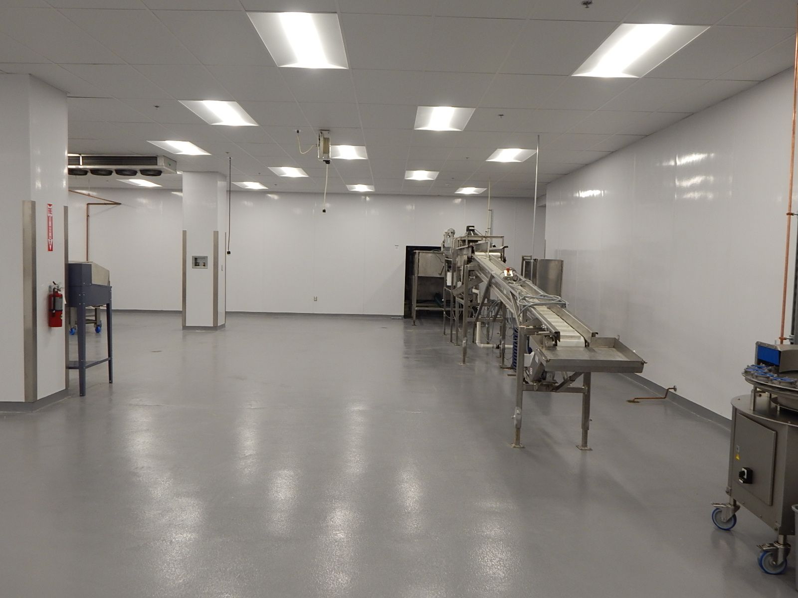 A bright, sterile industrial room with white walls, a grey floor, and a metal conveyor belt system for manufacturing.