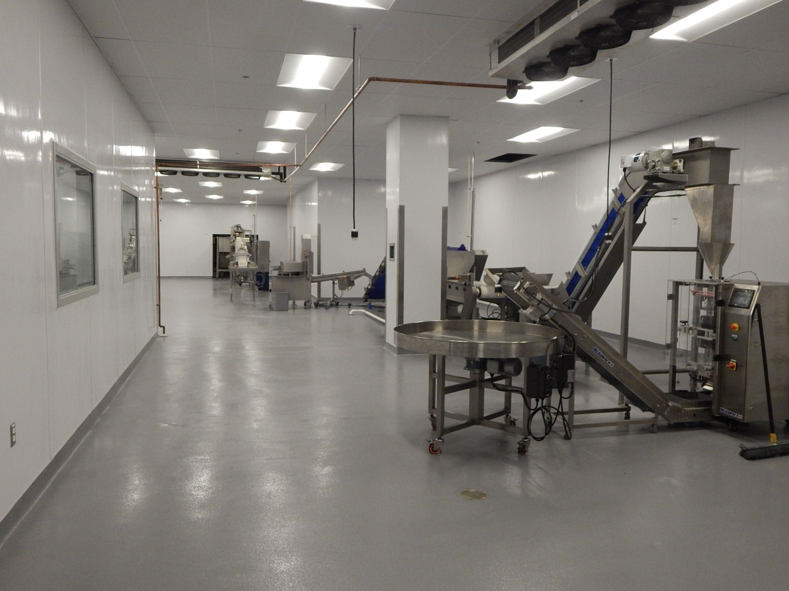 A clean, industrial facility with white walls, a gray floor, and stainless steel food processing and packaging machinery.