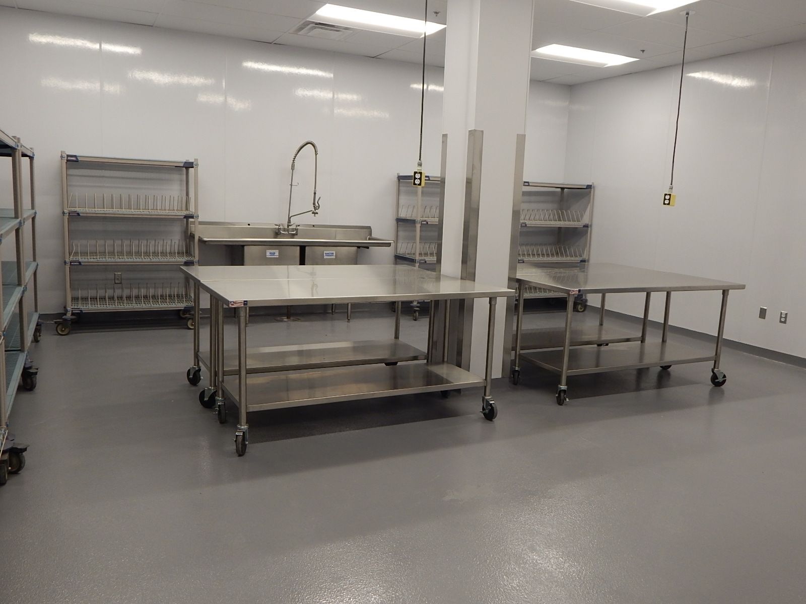 A clean, sterile laboratory or industrial workspace with stainless steel tables, shelving units, and a sink on white walls.