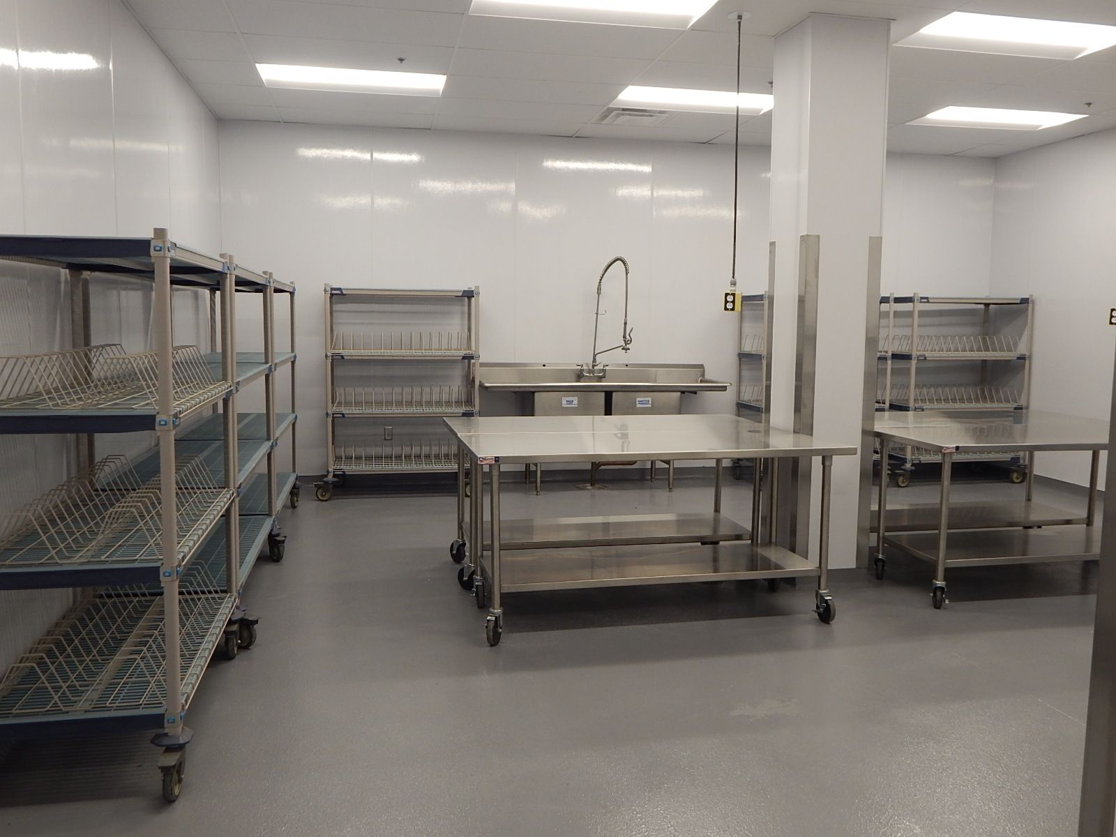 A brightly lit industrial kitchen or food prep area with metal shelving units, prep tables, and a sink on a grey floor.