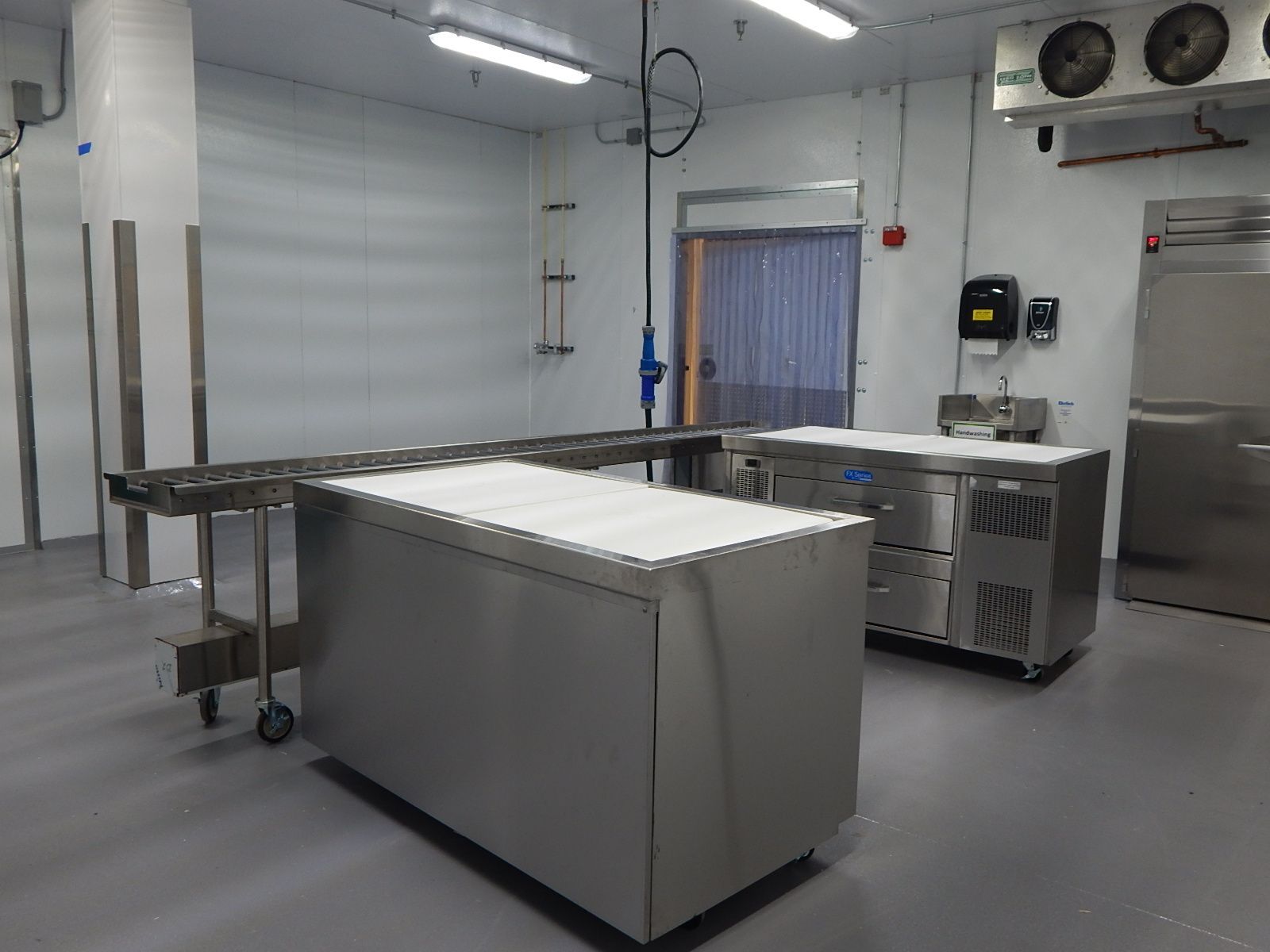 A clean, stainless-steel commercial food preparation area with metal tables and a conveyor system in a white room.