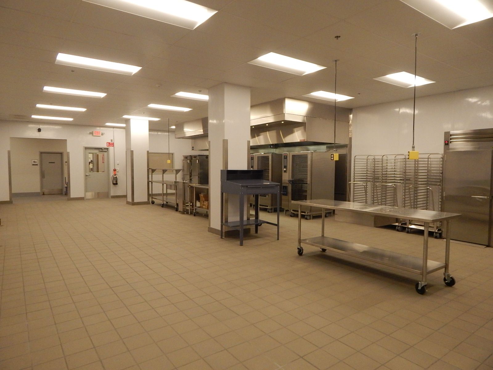 A brightly lit commercial kitchen with tiled floors, stainless steel workstations, shelving, and large appliances.