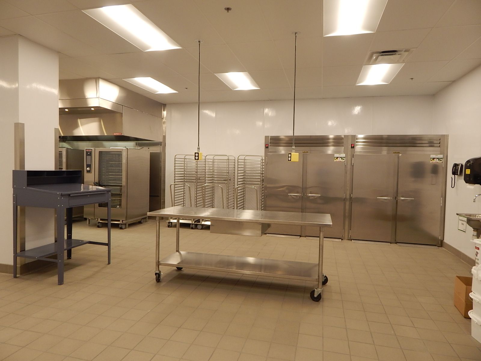 A commercial kitchen featuring stainless steel tables, refrigerators, and storage racks on a light-colored tile floor.