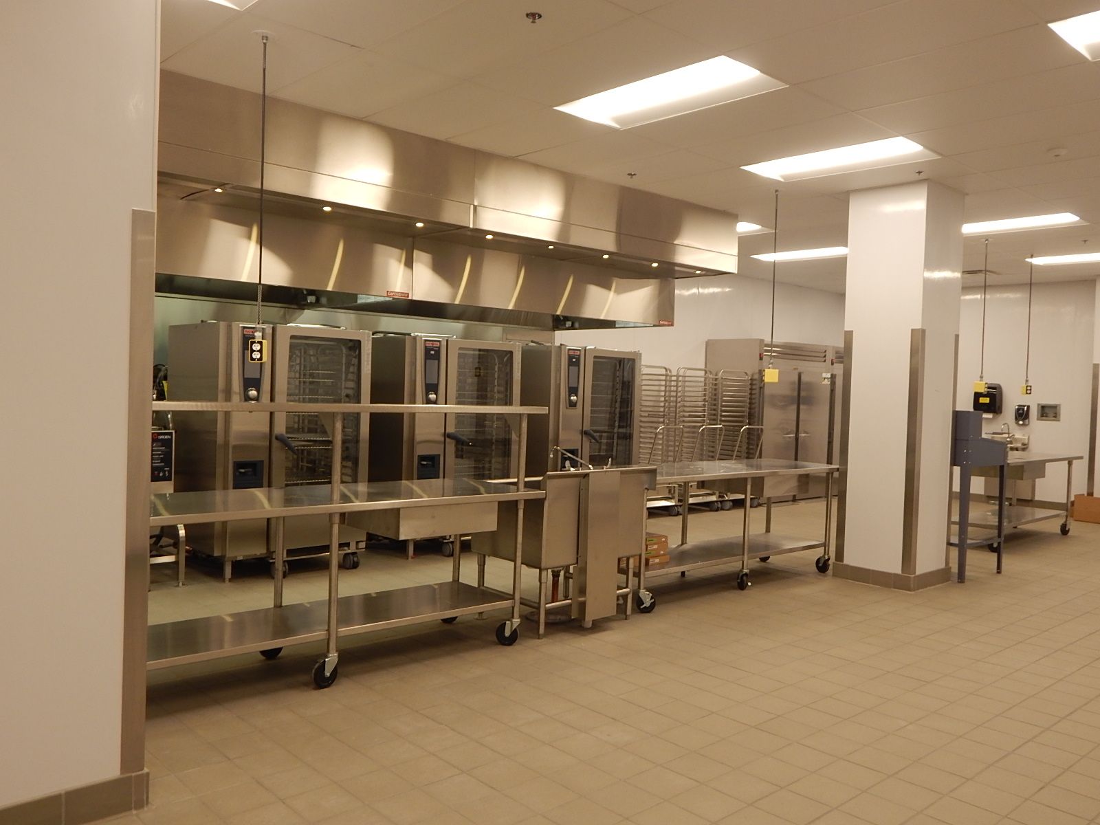 A clean, commercial kitchen with rows of stainless steel ovens, prep tables, and a large metal exhaust hood overhead.