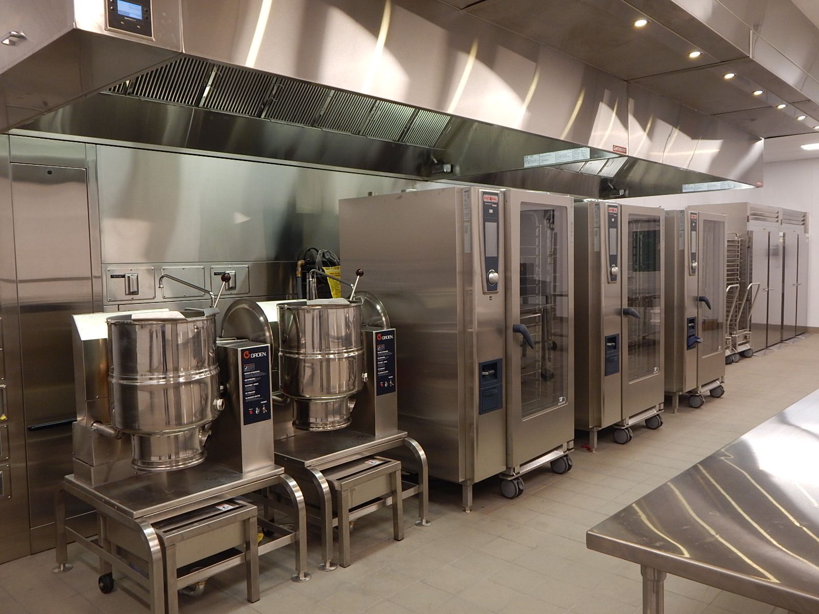 A row of stainless steel commercial kitchen equipment, including tilting skillets and ovens, under a large exhaust hood.
