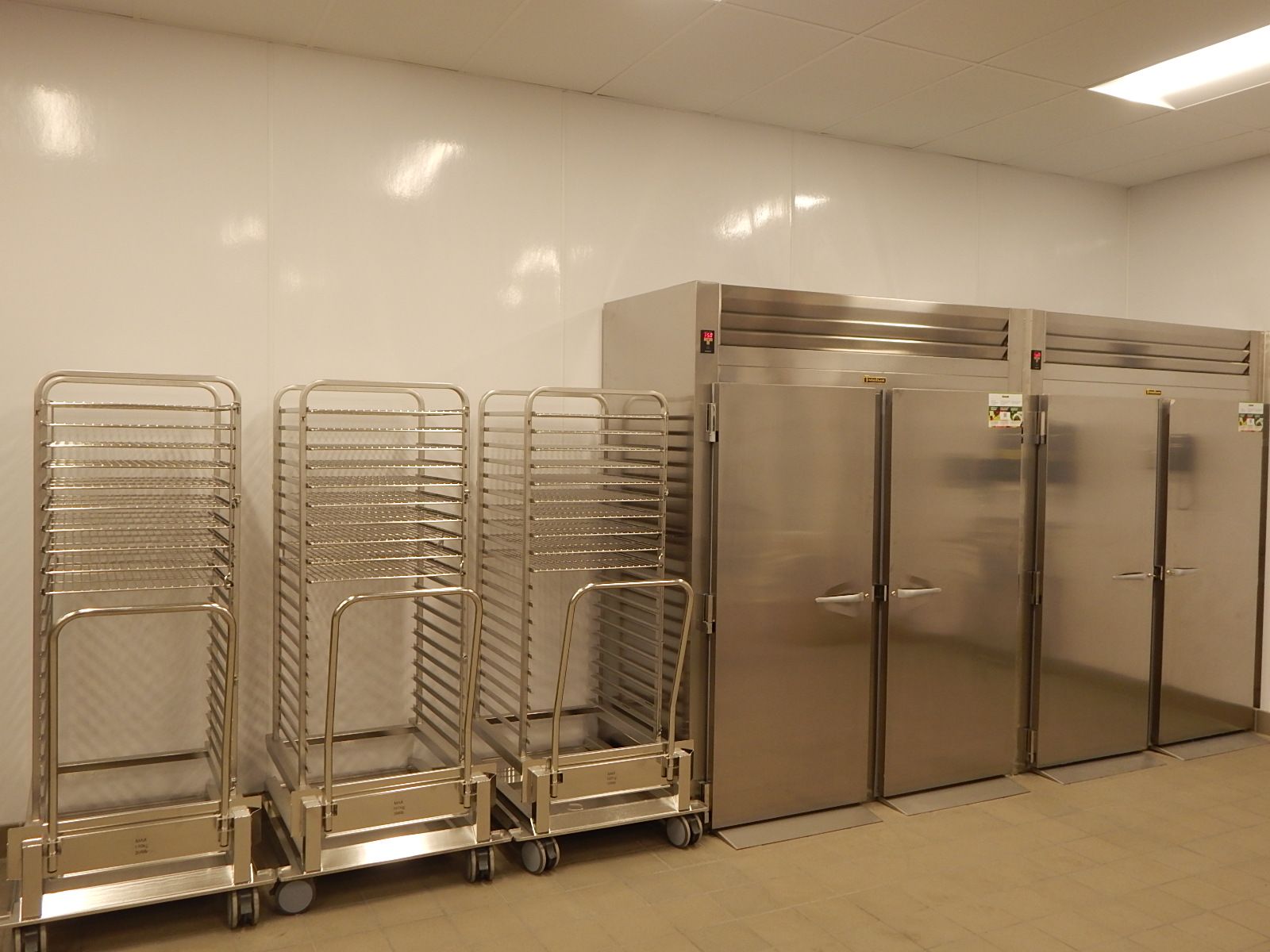 Three stainless steel rolling tray racks and two tall commercial refrigerators in a bright, clean professional kitchen.