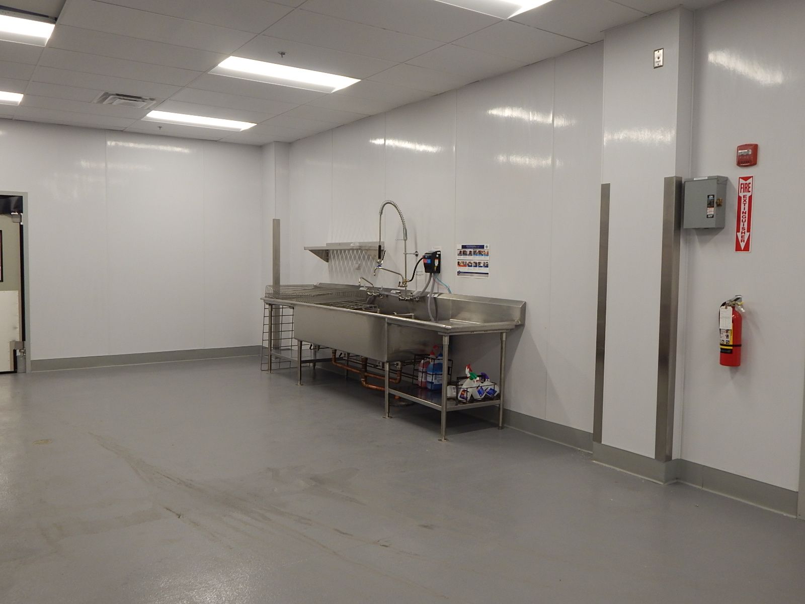 A clean, bright industrial room with white walls, a gray floor, a stainless steel sink station, and a wall fire extinguisher.