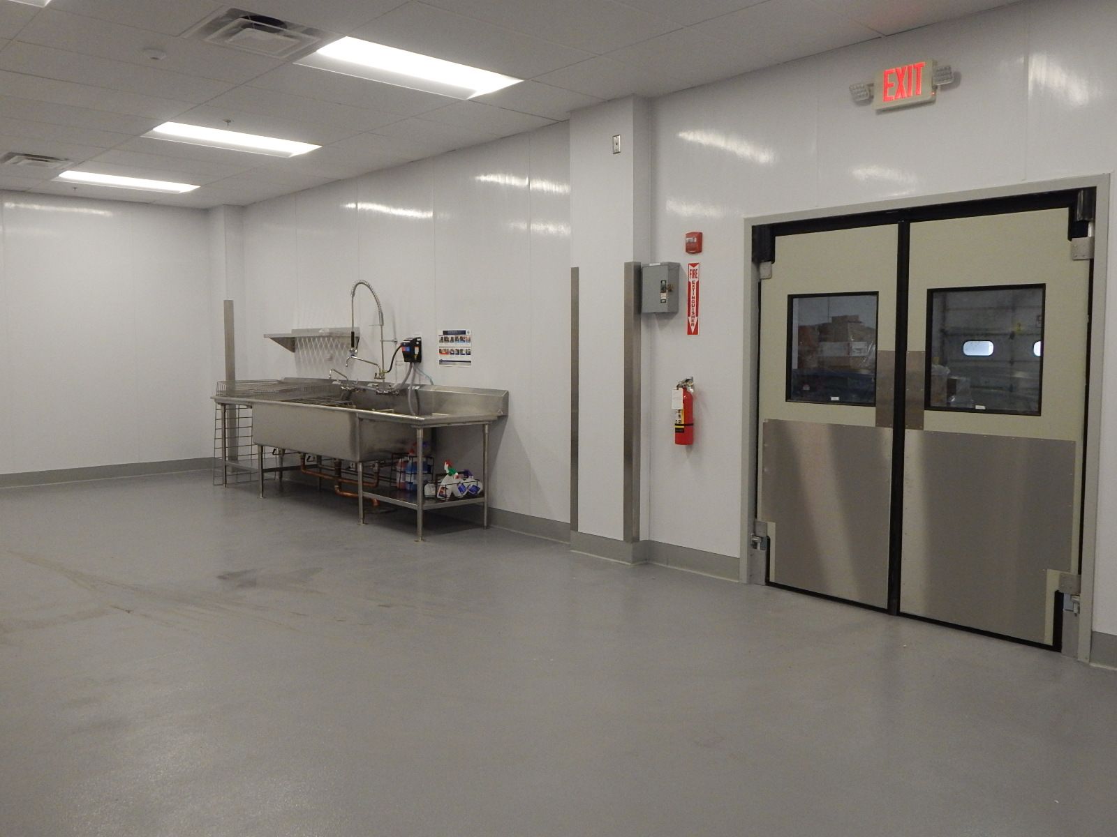 A clean, industrial-style room with white walls, a stainless steel sink station on the left, and double doors on the right.