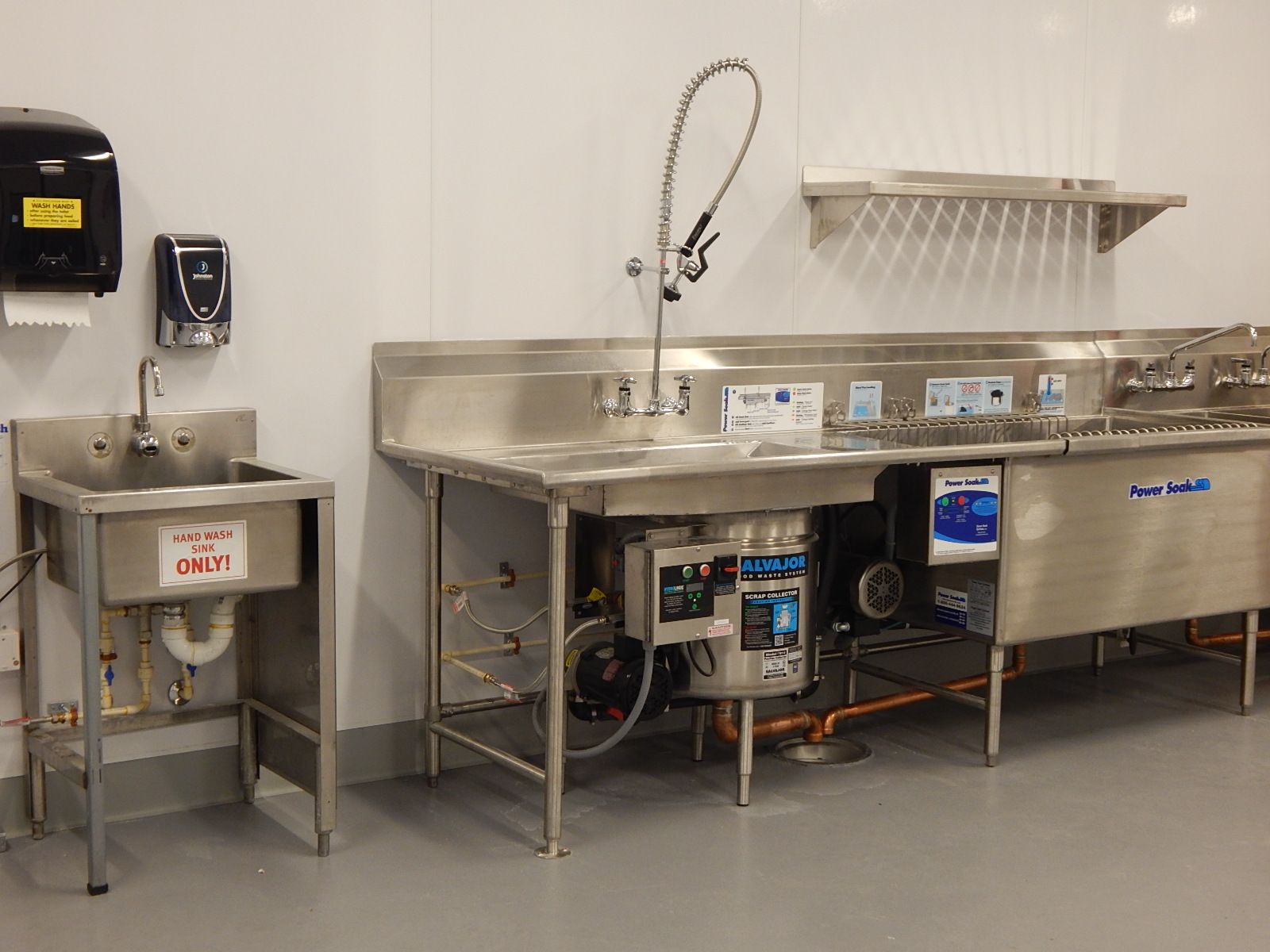A stainless steel commercial kitchen sink station with a pre-rinse sprayer and a wall-mounted hand-washing sink nearby.