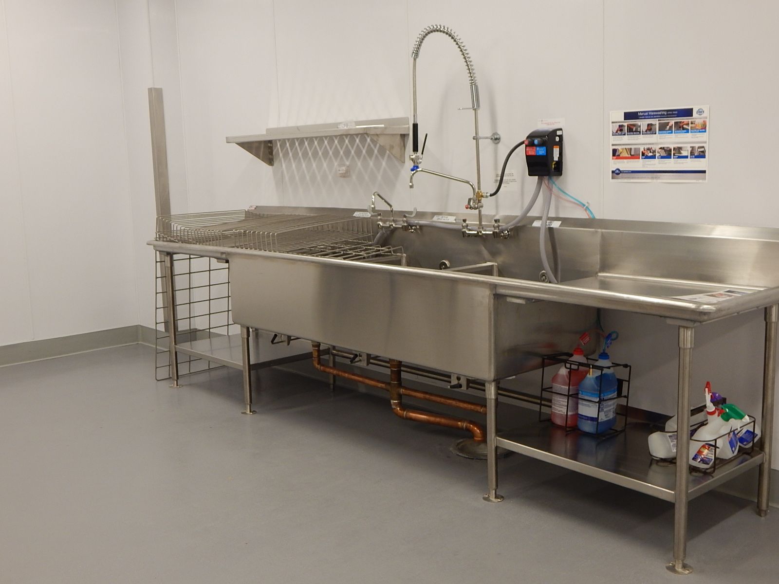 A stainless steel, three-compartment commercial sink with a spray faucet, drying rack, and cleaning supplies on a shelf.