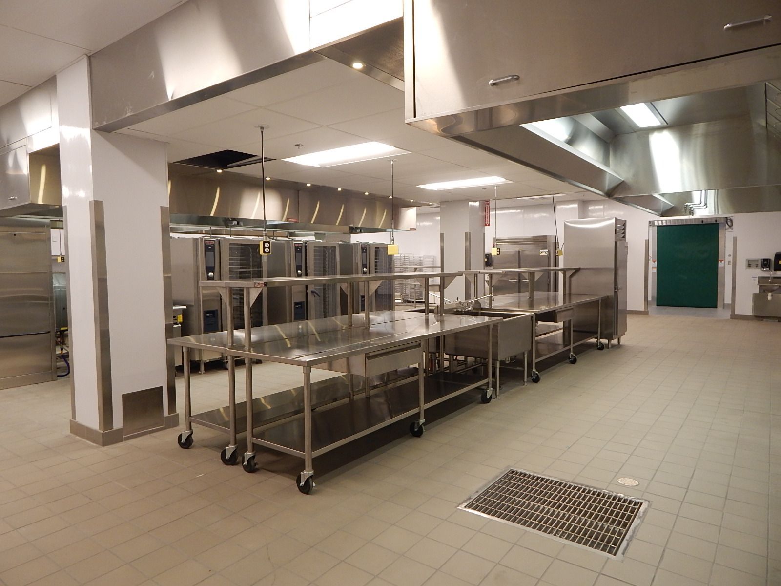 A wide, bright commercial kitchen featuring rows of stainless steel tables, appliances, and overhead vent hoods.