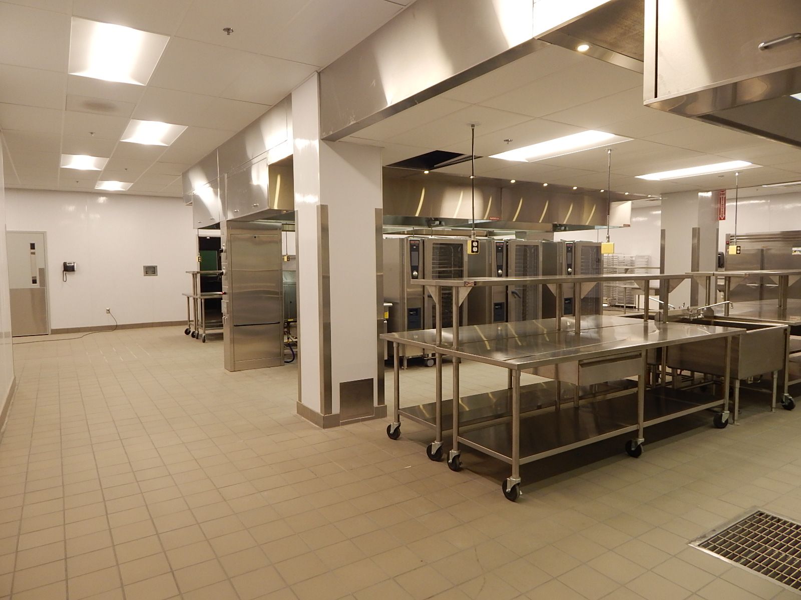 A spacious, modern industrial kitchen with stainless steel prep tables, cooking equipment, and white tiled floors.