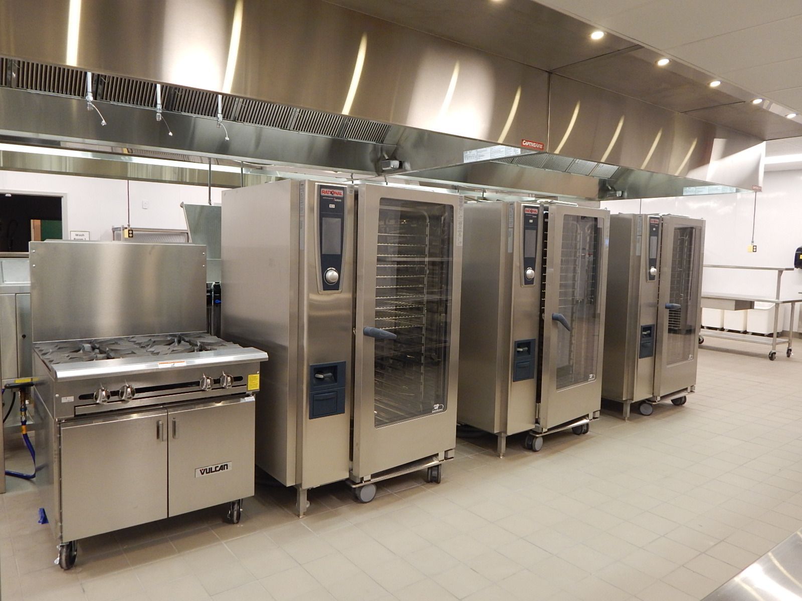 A commercial kitchen with a stainless steel stove next to three tall, professional stainless steel convection ovens.