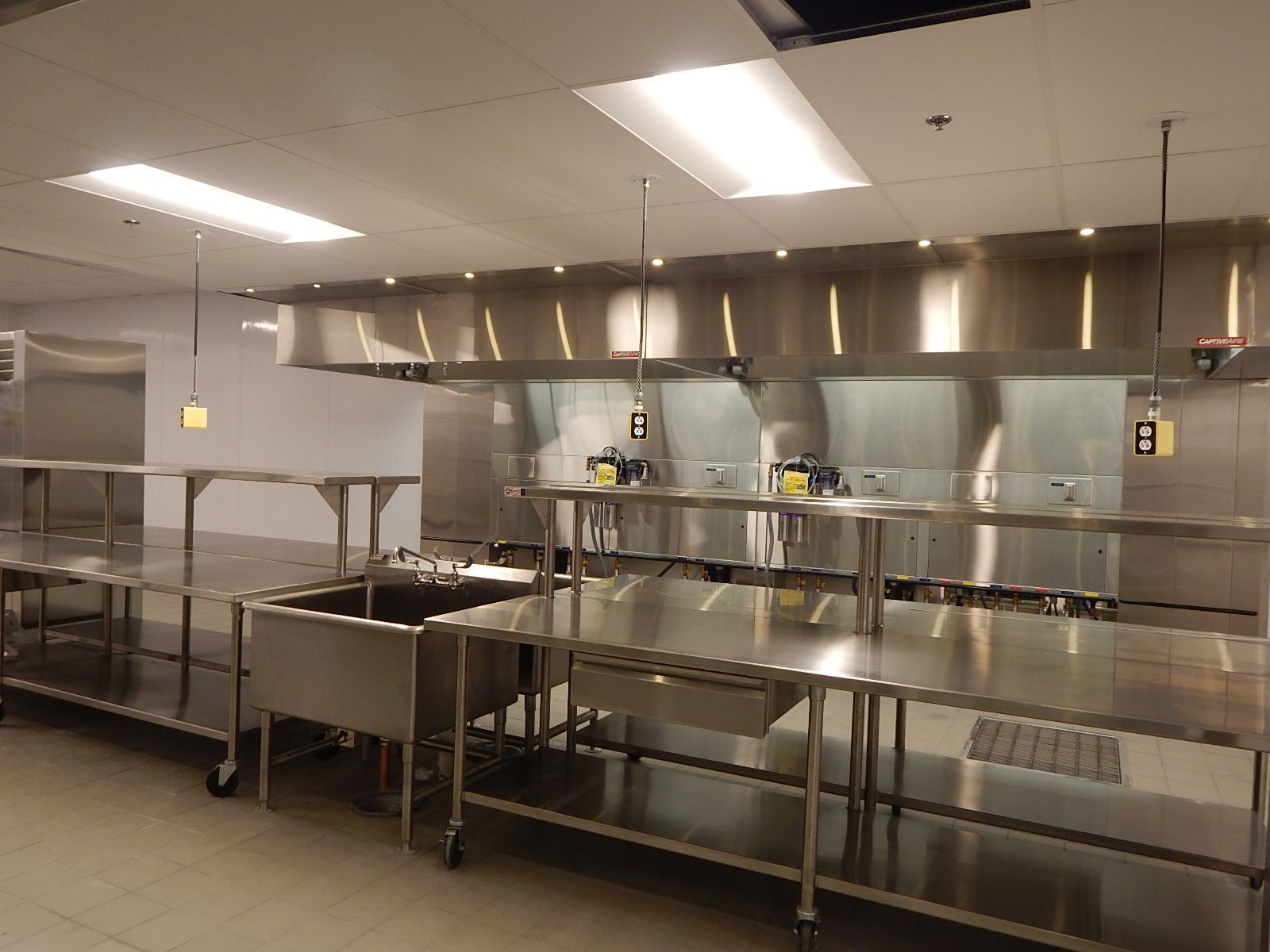 Commercial kitchen interior with stainless steel prep tables, sinks, and large overhead ventilation hoods.