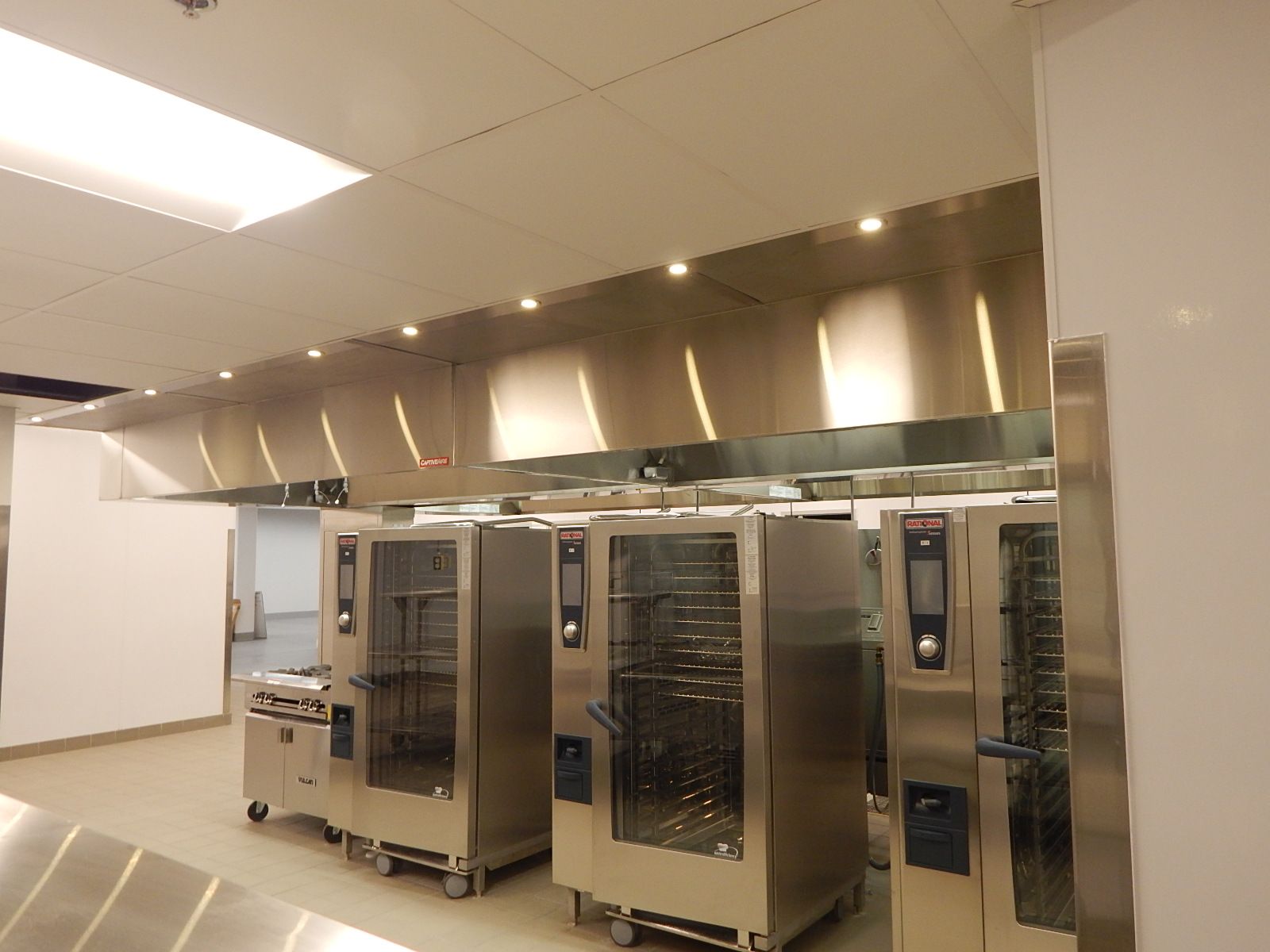 A commercial kitchen featuring three tall stainless steel Rational ovens beneath a large, well-lit metallic vent hood.