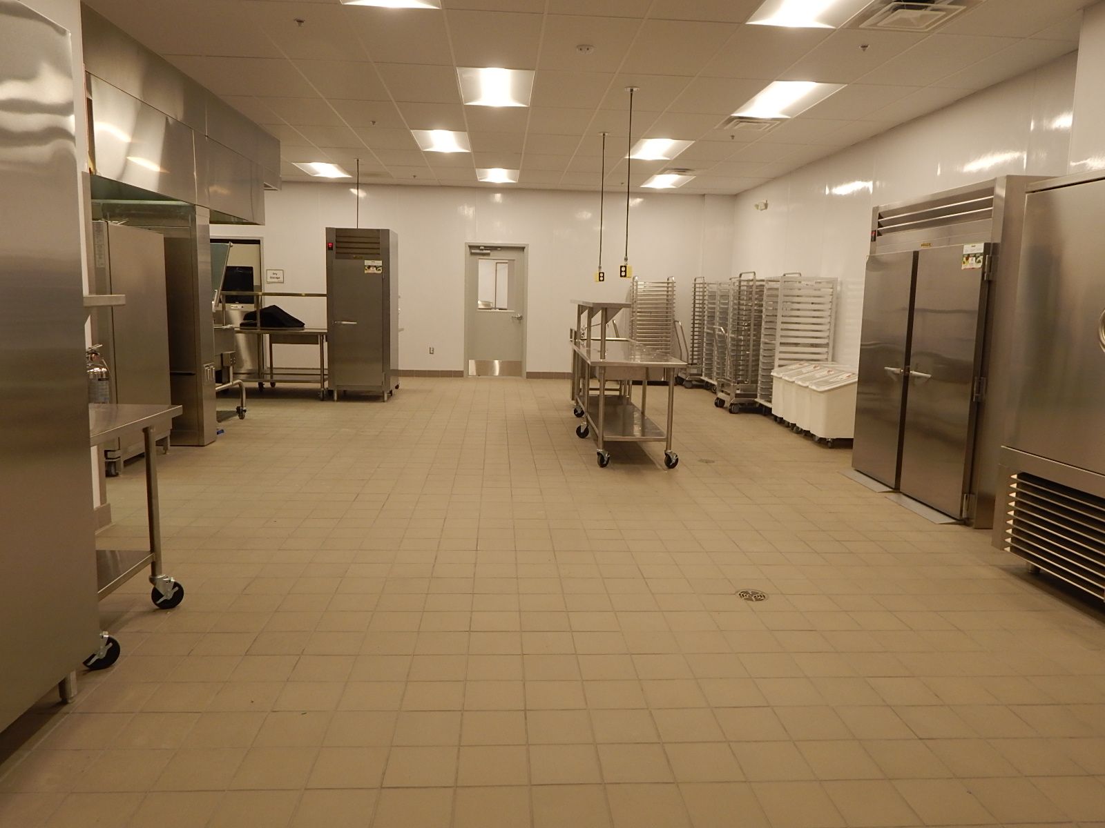 A commercial kitchen featuring stainless steel appliances, prep tables on wheels, and wire racks on a light tile floor.