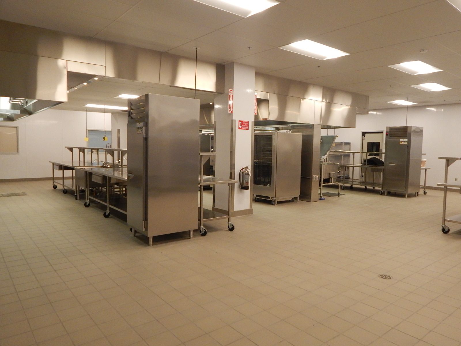 A bright, clean, commercial kitchen with stainless steel appliances, prep tables, and light-colored tile floors.
