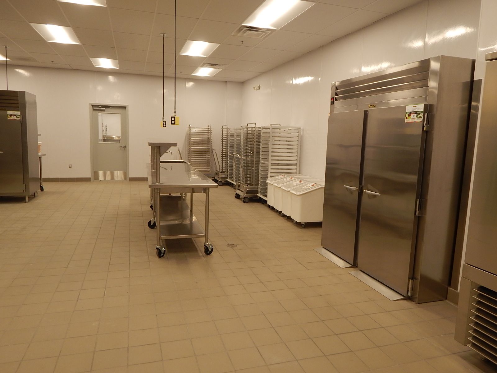 A commercial kitchen with stainless steel refrigerators, rolling metal carts, a prep table, and light tile flooring.