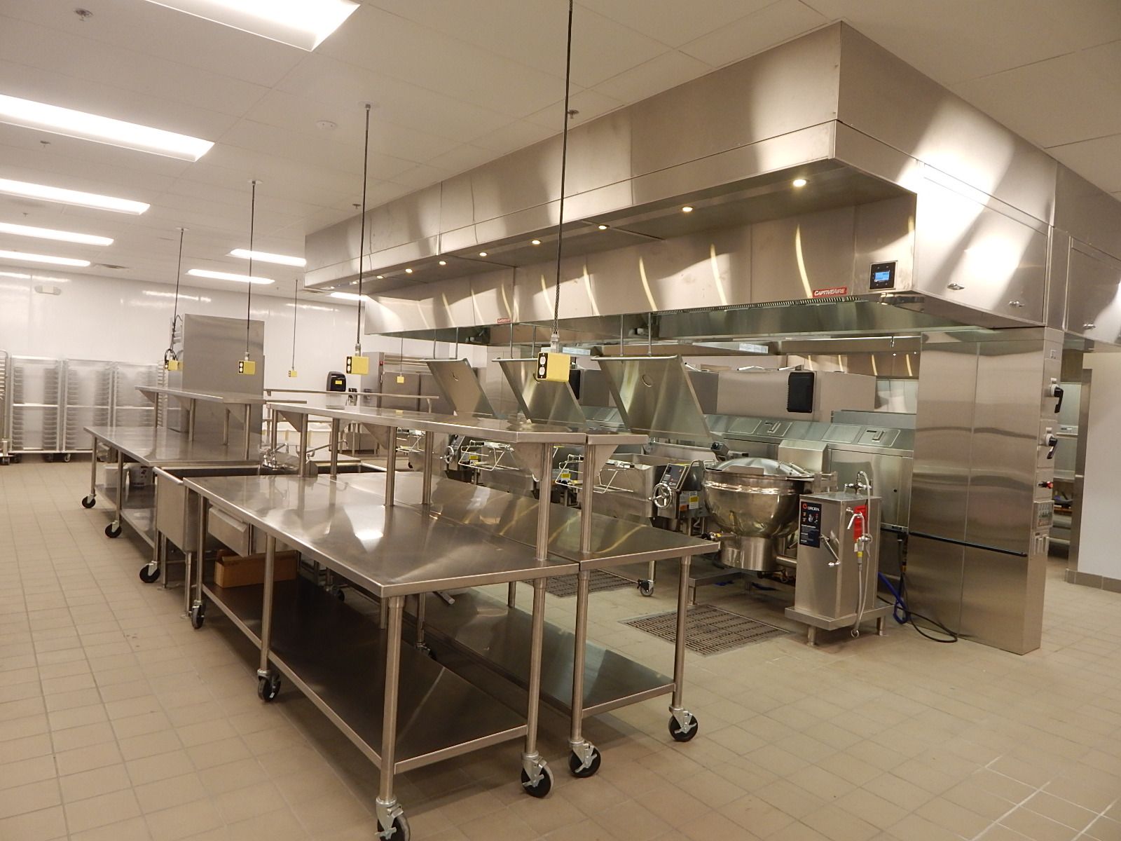 A stainless steel commercial kitchen featuring rows of metal prep tables and large cooking equipment under vent hoods.