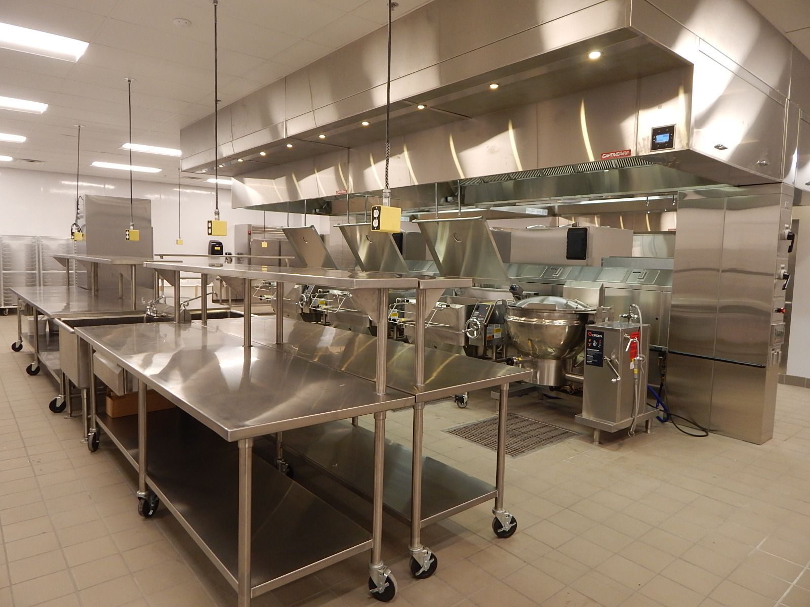 A clean, modern commercial kitchen with stainless steel prep tables, cooking equipment, and a large metal exhaust hood.