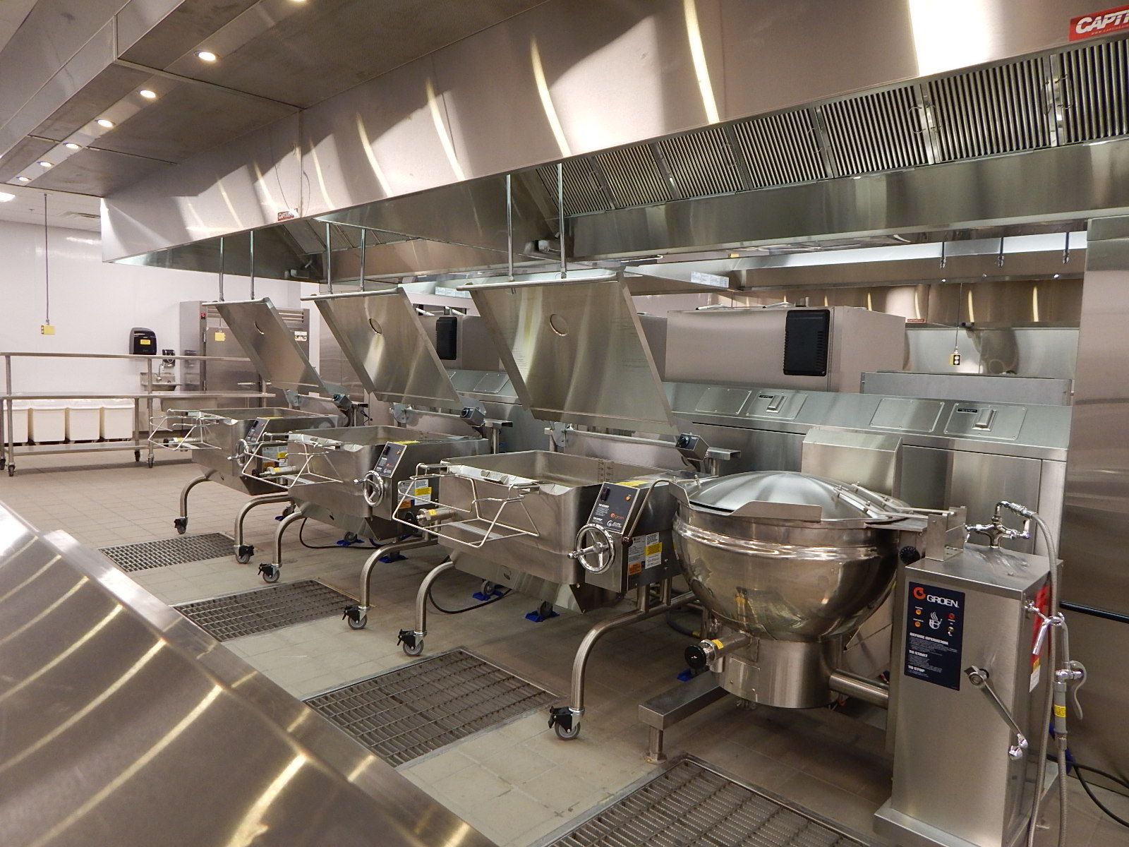 A row of industrial stainless steel cooking kettles and tilting skillets under a large ventilation hood in a kitchen.